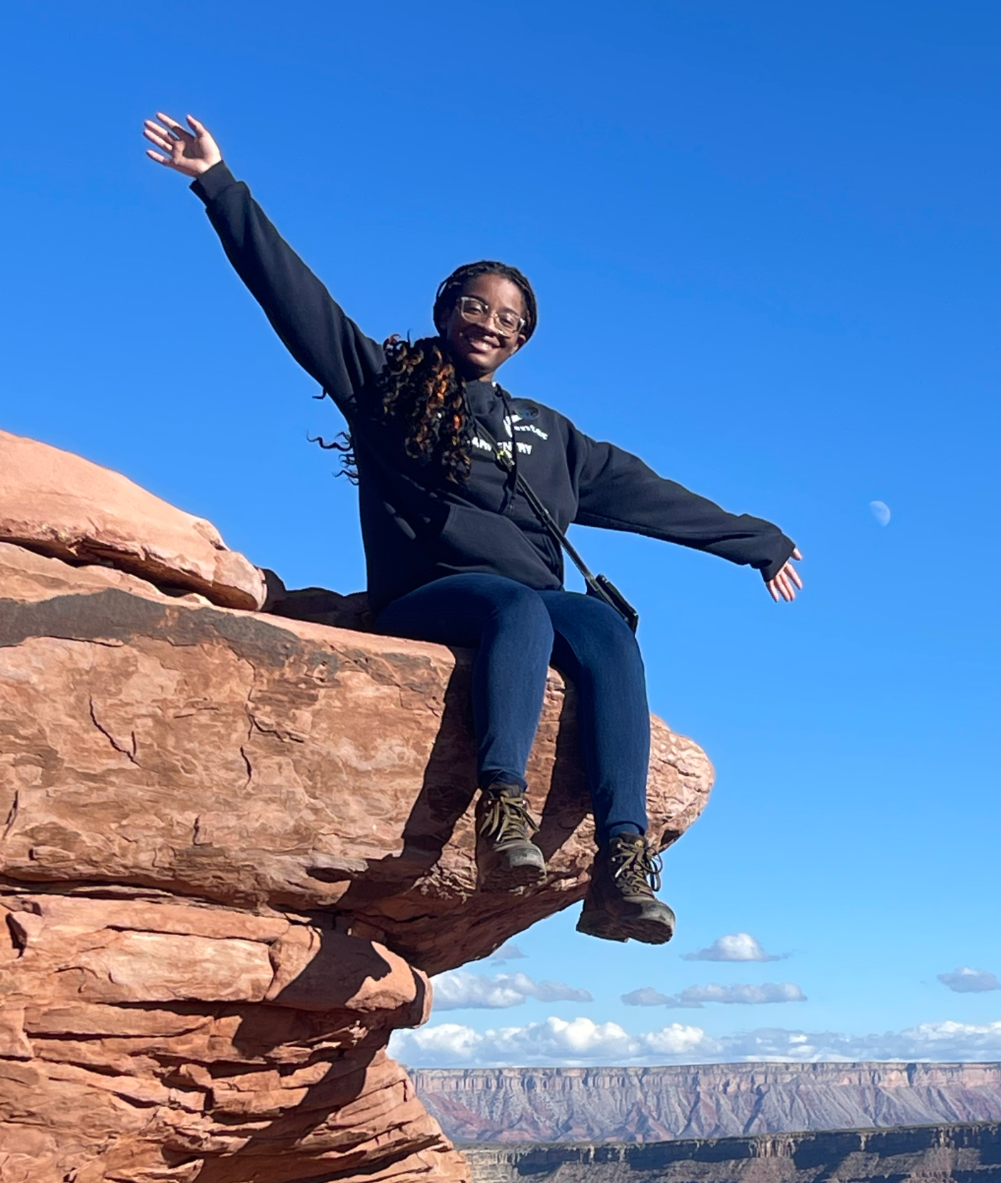 Laila Wilson sits atop the edge of a stone formation with her legs over the ledge and arms out wide. A deep canyon in the background and bright blue skies.